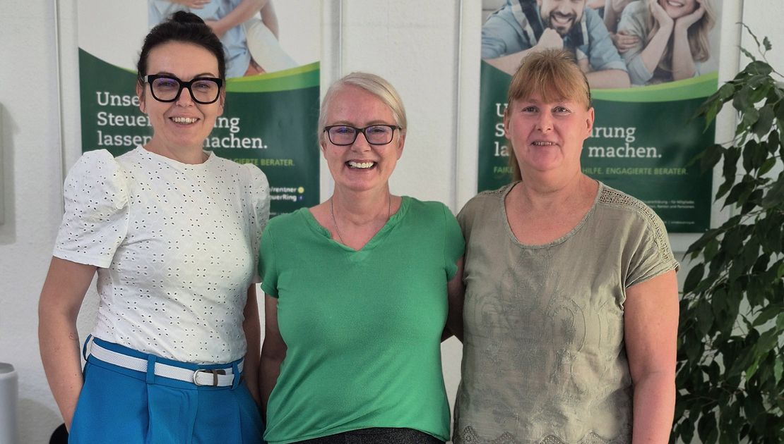 Drei Frauen stehen nebeneinander in einem Büro und schauen in die Kamera. Links trägt eine Frau mit dunklen Haaren und Brille eine weiße Bluse, eine blaue Hose und weiße Schuhe. In der Mitte steht eine blonde Frau mit Brille, grünem Oberteil und dunkler Hose. Rechts trägt eine Frau ein graugrünes Oberteil, eine helle weite Hose und helle Schuhe. Hinter ihnen hängen zwei grüne Werbeplakate des Steuerrings mit Bildern von Familien; rechts steht eine große Zimmerpflanze.