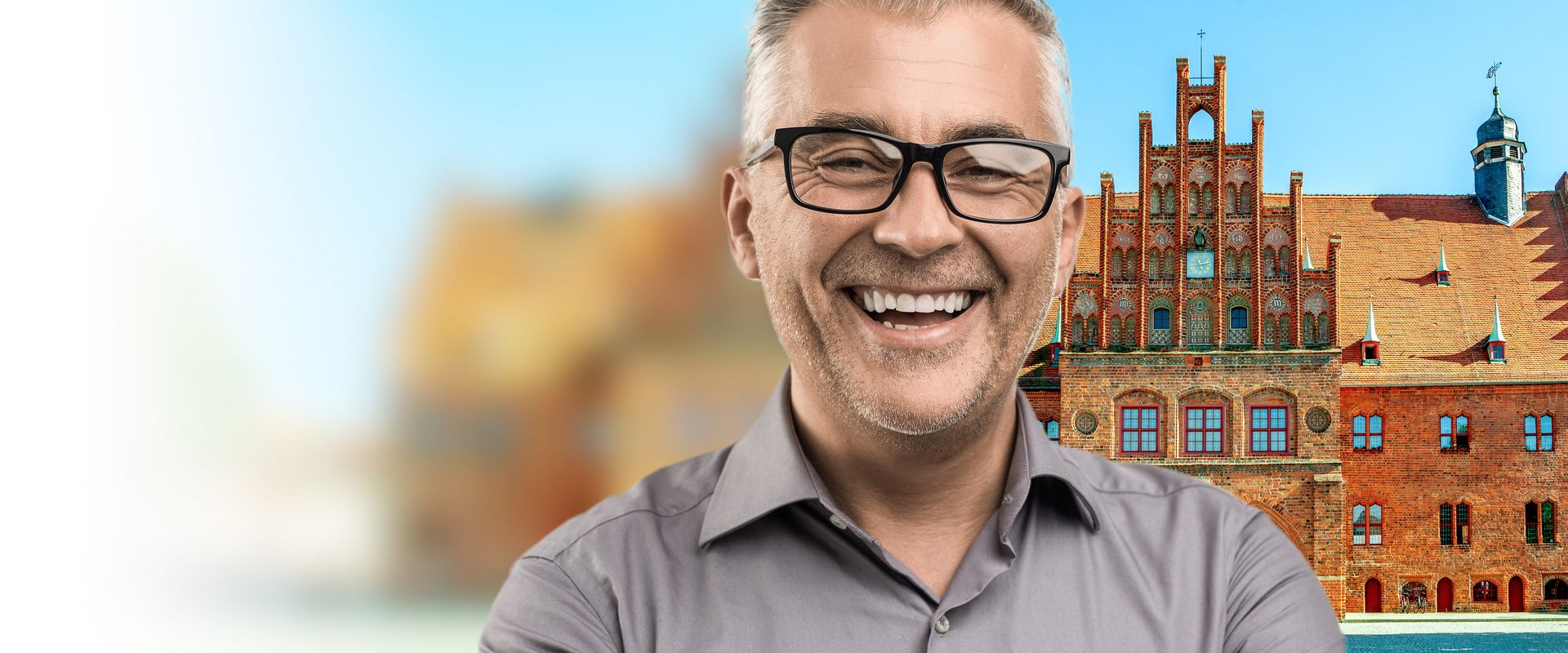 Ein Mann mit Brille lächelt in die Kamera, im Hintergrund steht ein großes historisches Backsteingebäude. Der Mann ist im Vordergrund von der Brust aufwärts zu sehen und trägt ein graues Hemd, hinter ihm ist ein reich verziertes Gebäude mit vielen Fenstern, einem Giebel und einem Turm, der linke Bildbereich ist unscharf und hell.