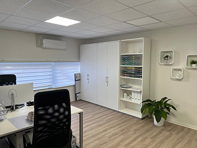 Ein helles Büro mit zwei Arbeitsplätzen, einem großen weißen Schrank und einem Regal rechts. Helle Wände, Fenster mit Rollo, offene Regale mit Unterlagen und Deko sowie eine Pflanze am Boden.