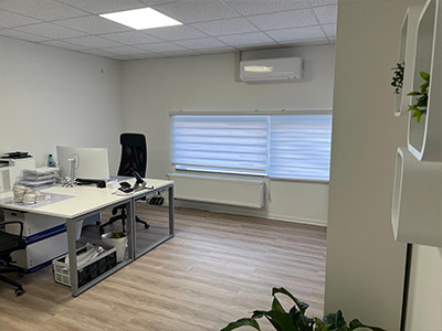 Ein helles Büro mit einem einzelnen Arbeitsplatz, einem großen Fenster mit Rollo und hellem Boden. Links steht ein Schreibtisch mit Monitor, Stuhl und Bürobedarf, rechts sind Wandregale mit Pflanzen zu sehen.