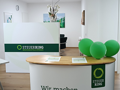 Man sieht einen hellen Raum, im Vordergrund eine Art Kommode und im Hintergrund eine Empfangstheke, beide in weiß mit Steuerring-Logo