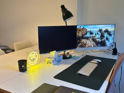 Moderner Arbeitsplatz mit zwei Monitoren auf einem weißen Schreibtisch, einer Schreibtischlampe, Tastatur, Maus und einer leuchtenden Smiley-Lampe; auf einem Bildschirm ist eine Küstenlandschaft zu sehen.