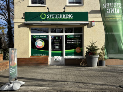 Eingangsbereich eines Steuerbüros mit grüner Beschilderung „Steuerring“, Glastür mit Informationsaufklebern, Pflanze im Topf daneben und gepflastertem Gehweg davor.