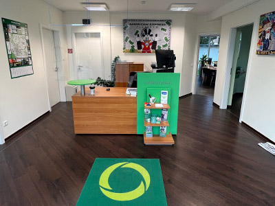 Empfangsbereich im Steuerring-Büro Ibbenbüren Ein Weitinkelbild eines Empfangsbereichs mit grünem Teppich auf dem das Steuerring-Logo prangt. Eine Holztheke, brauner Laminatboden und weiße helle Wände. An den Wänden sind unter anderem Bilder von Mickey Mouse.