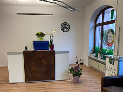 Ein modern eingerichtetes Büro mit einem Empfangstresen im Vordergrund. Der Tresen ist weiß mit einer braunen Holzverkleidung in der Mitte. Links vom Tresen steht ein blauer Aktenschrank mit einer Orchidee darauf. Auf der rechten Seite des Raumes steht ein großes Fenster mit Blick nach draußen, welches mit grünen Stickern beklebt ist. Neben dem Fenster steht ein kleiner Tisch mit Werbematerialien. An der Wand hinter dem Empfangstresen hängt eine Uhr. Der Boden besteht aus hellem Holz.