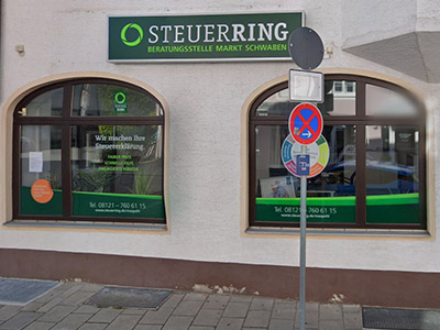 Das Bild zeigt die Außenansicht eines Gebäudes mit dem Schild "STEUERRING Beratungsstelle Markt Schwaben". Es gibt zwei große Fenster mit abgerundeten oberen Ecken, durch die man in das Büro schauen kann. Die Fenster sind mit Aufklebern dekoriert, die auf die Dienstleistungen des Steuerberatungsbüros hinweisen. Vor einem Fenster steht ein Schild mit einem Halteverbotsschild und einem Parkverbotsschild darunter. Der Bürgersteig vor dem Gebäude ist gepflastert.