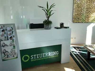 Heller Eingangsbereich des Steuerring-Büros in Rosenheim. Der Thresen ist weiß und hat ein grünes Panel in Steuerring-Farben mit dem hellgrünen Logo und weißen Schriftzugdarauf. Man sieht noch ein Stück eines Teppichs und wie die Sonne einstrahlt.