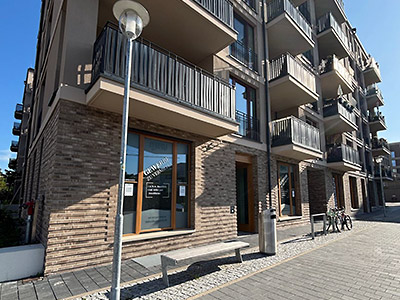 Moderne Wohn- und Geschäftsgebäude mit Balkonen aus hellem Backstein. Im Erdgeschoss ist ein Schaufenster mit Beschriftung zu sehen, davor stehen eine Bank, ein Mülleimer und Fahrräder auf einem gepflasterten Gehweg.