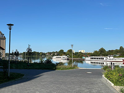 Blick auf einen ruhigen Kanal unter blauem Himmel. Mehrere weiße Ausflugsboote liegen am Ufer, umgeben von grünen Bäumen. Im Vordergrund führt ein gepflasterter Weg mit Straßenlaternen zum Wasser.